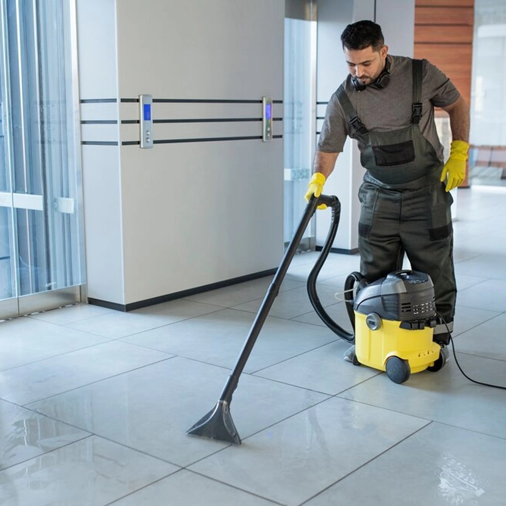 Worker in coveralls and yellow gloves using a wet/dry vacuum on a tile floor in a bright, empty building